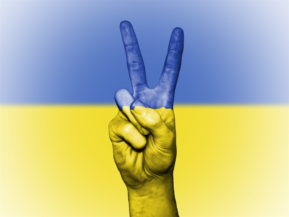 ID: Hand with index and middle finger raised in peace sign, overlaid with colors of the Ukraine flag, blue on top half, yellow on bottom.